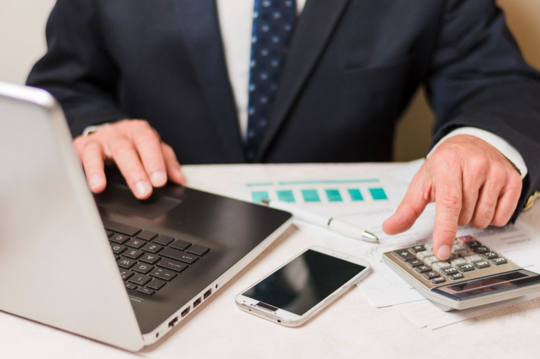 Person in suit using laptop, calculator, and smartphone at a desk with graphs.
