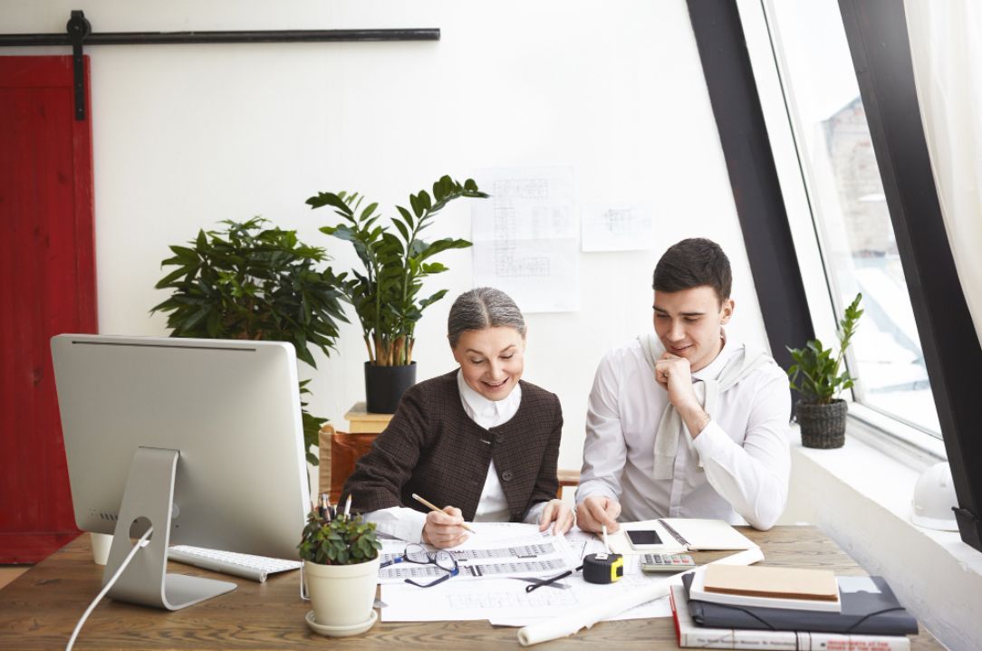 Woman and man at desk, reviewing documents. Woman points at paper, man looks thoughtful, room with plants.