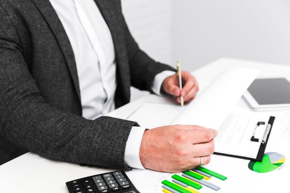 A person in a gray blazer writes on paper at a desk with a calculator, documents, and a small pie chart.