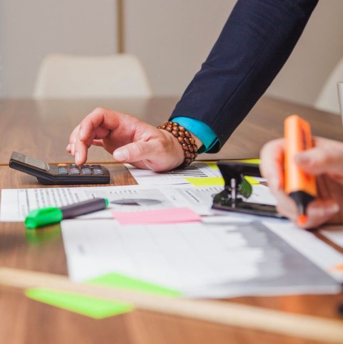 A person in a blue shirt and blazer uses a calculator on a desk filled with business charts, markers, and a highlighter.