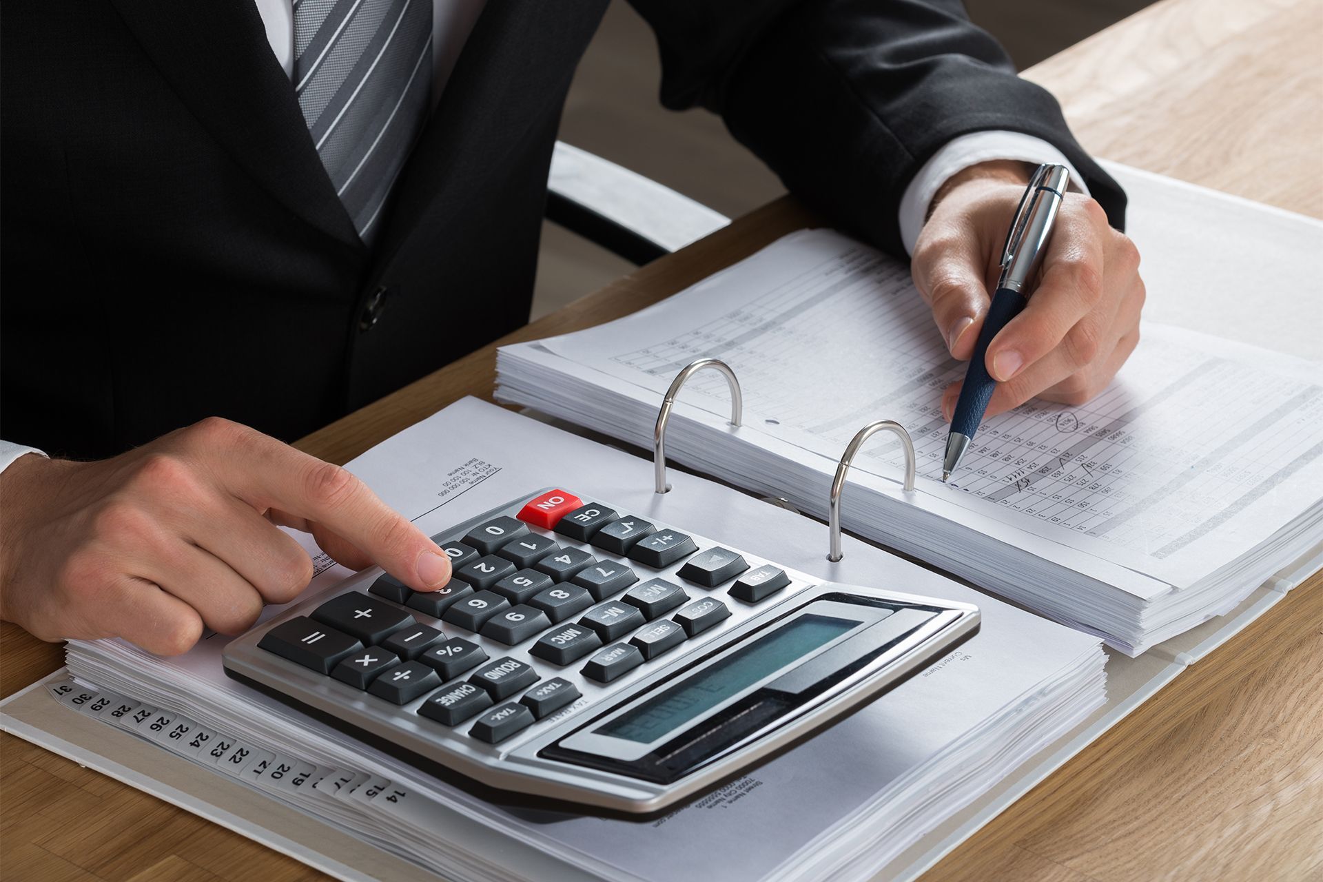 Person in a suit calculating with calculator and writing on papers at a desk.