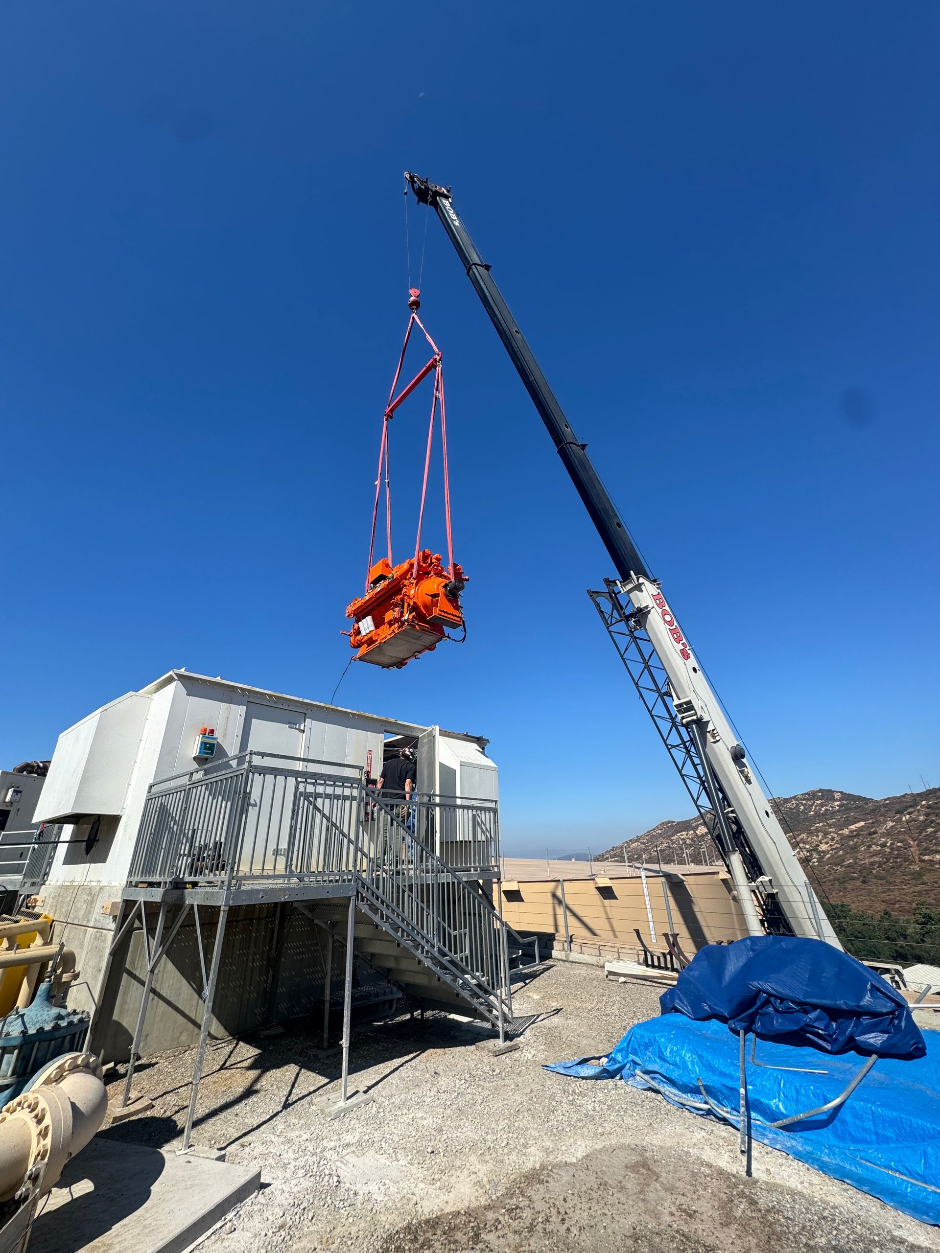 A crane lifts an orange-colored piece of machinery near a white building under a clear blue sky.
