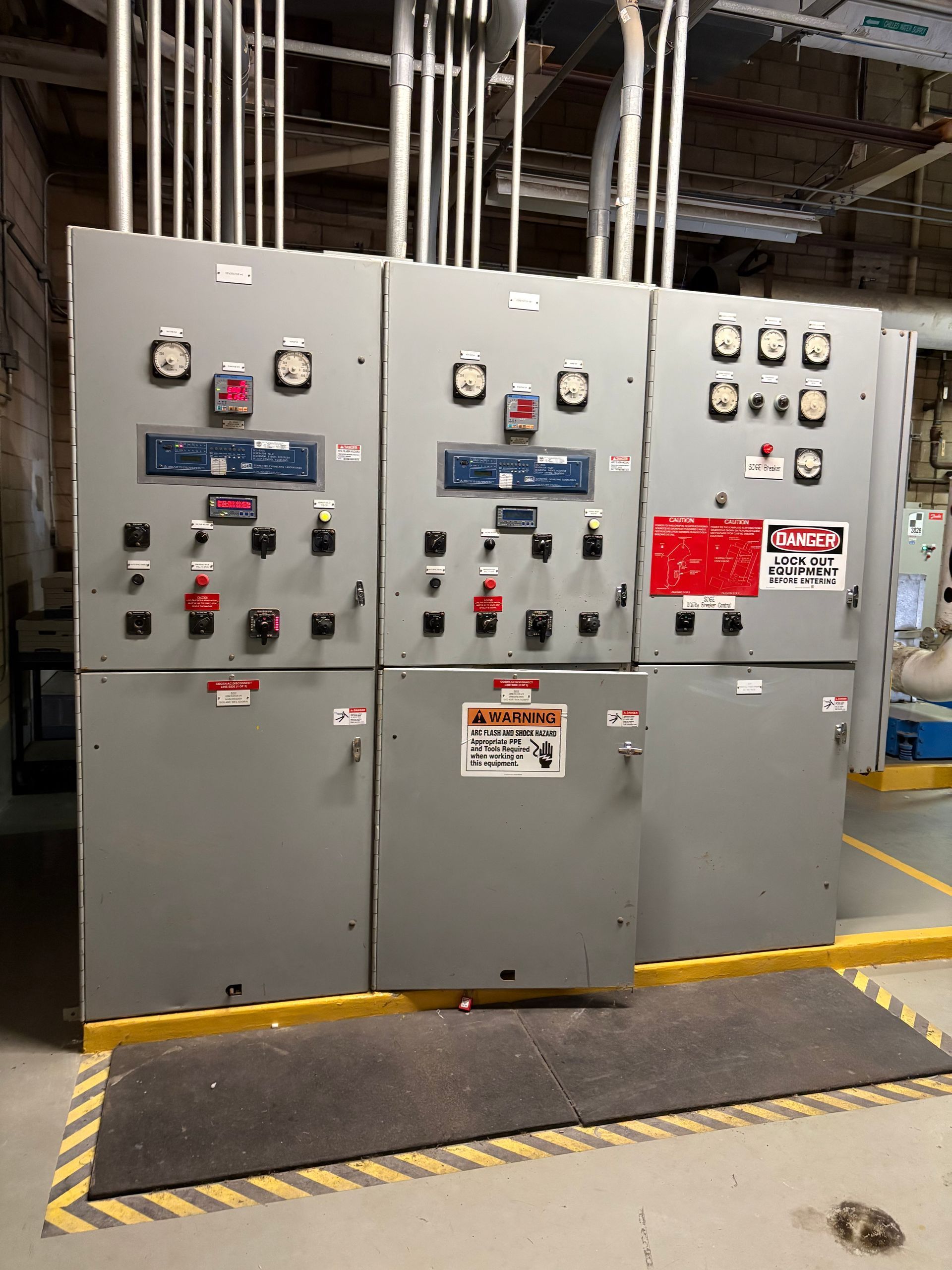 Electrical control panels in a gray industrial setting, with warning signs and conduits above.