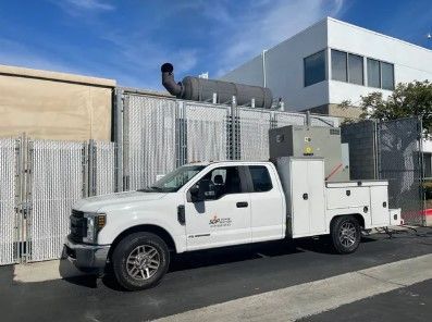 White work truck parked outside a building with a fence, equipment, and a blue sky.