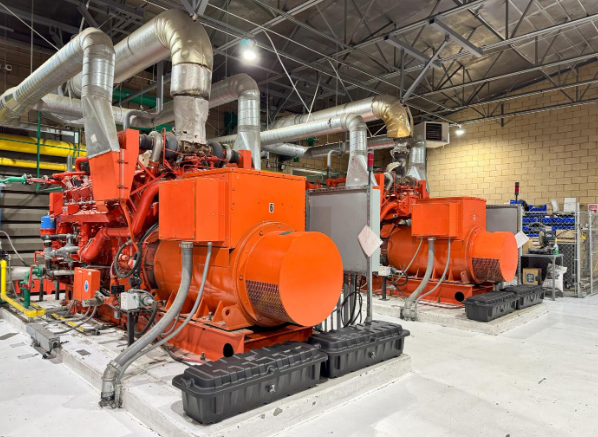 Two large orange generators in an industrial setting with silver piping overhead.