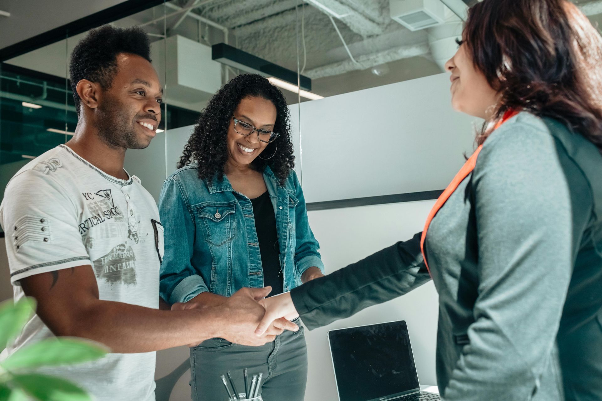 Two people shake hands in an office, while a third person looks on and smiles.