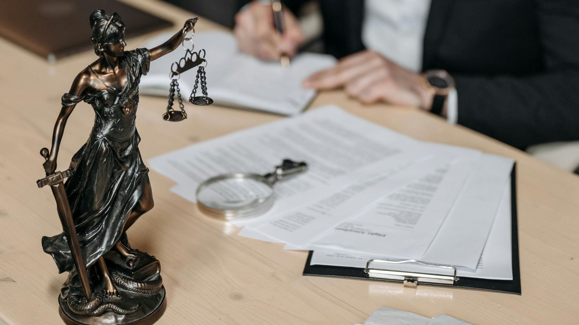 A statue of Lady Justice stands on a wooden desk next to legal documents, a magnifying glass, and a person writing.