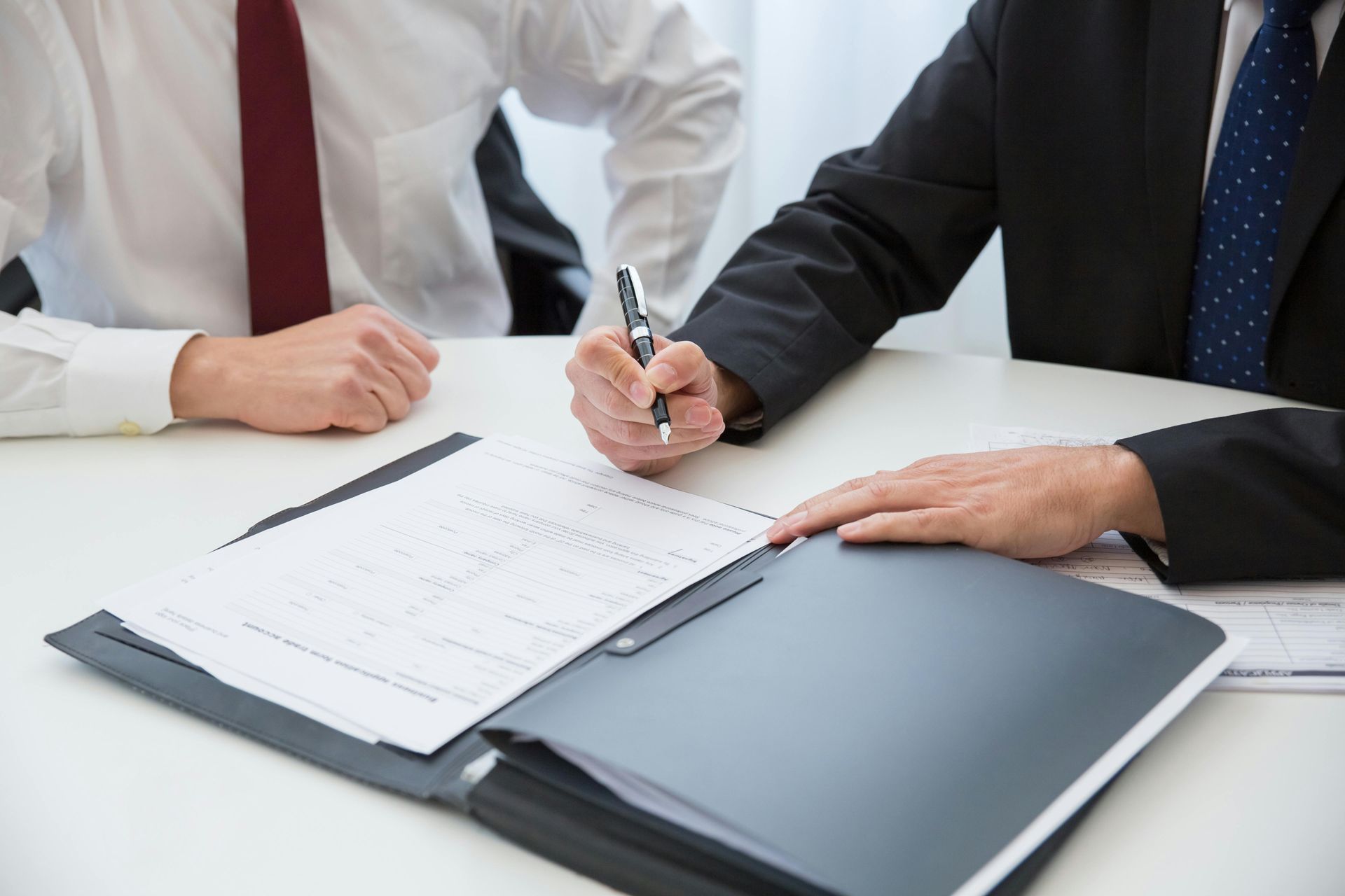 Two business professionals in formal attire sit at a desk, with one preparing to sign a document in a folder.