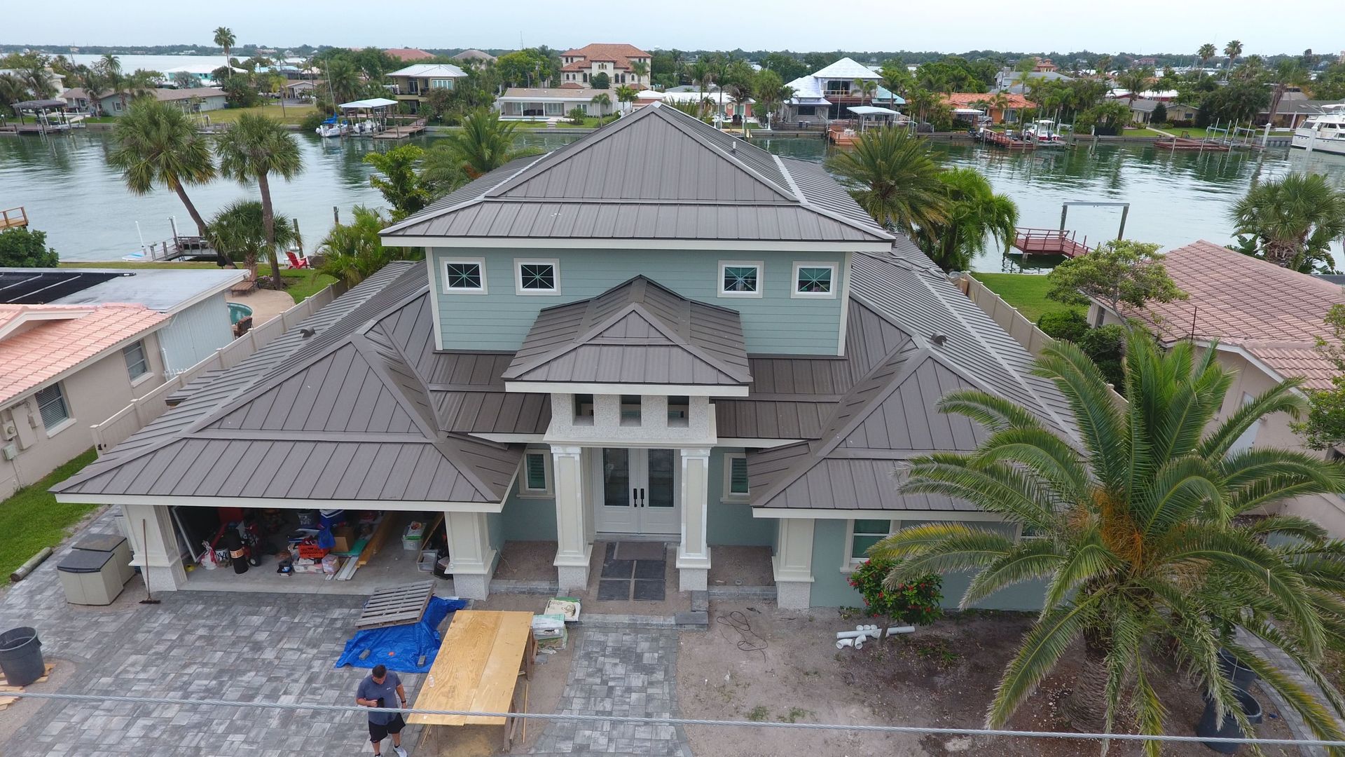 A light blue house with a dark gray roof sits on a waterfront, construction in progress, person outside.
