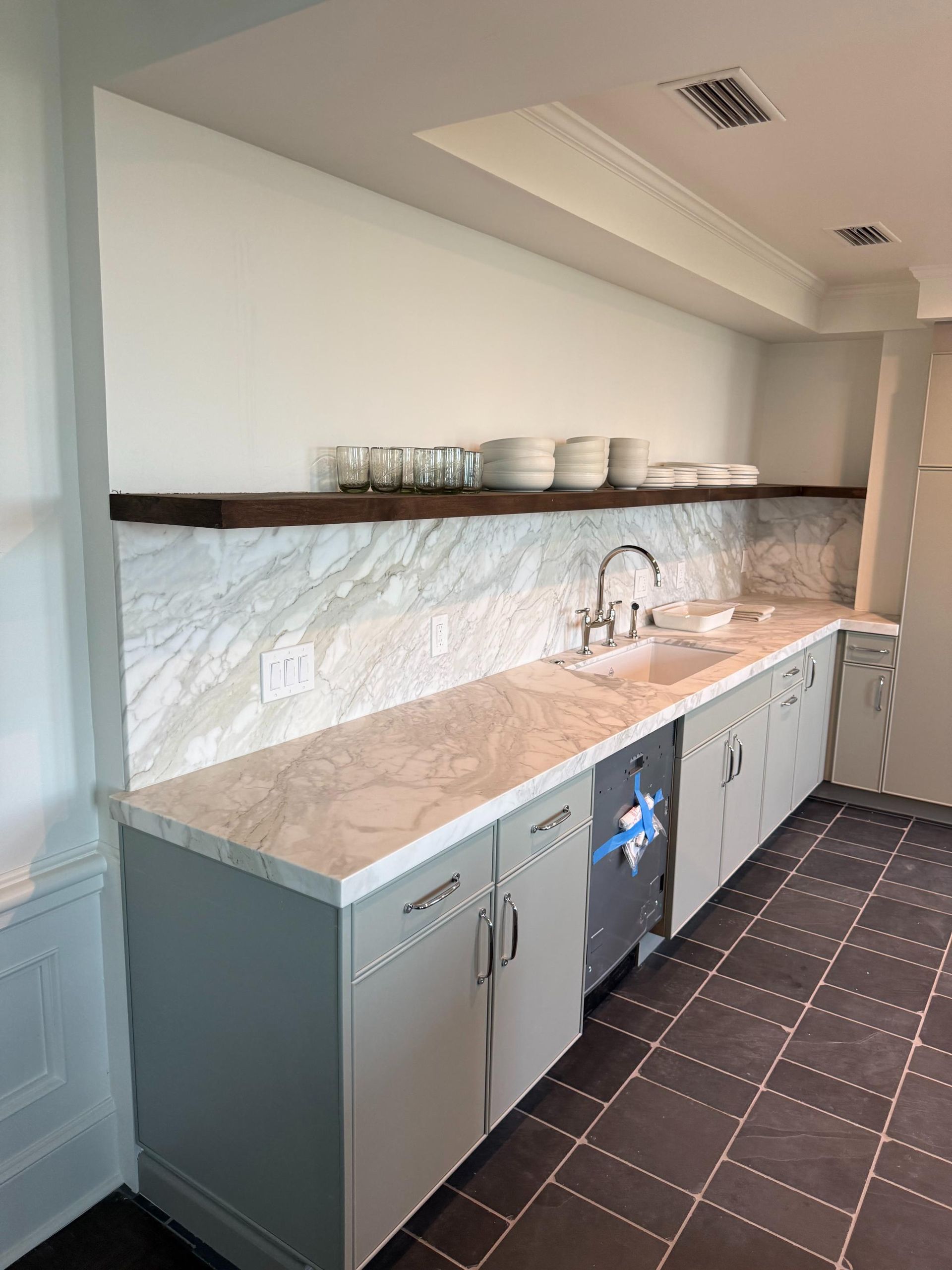 Kitchen with marble backsplash, gray cabinets, stainless steel sink, and dishes on a shelf.