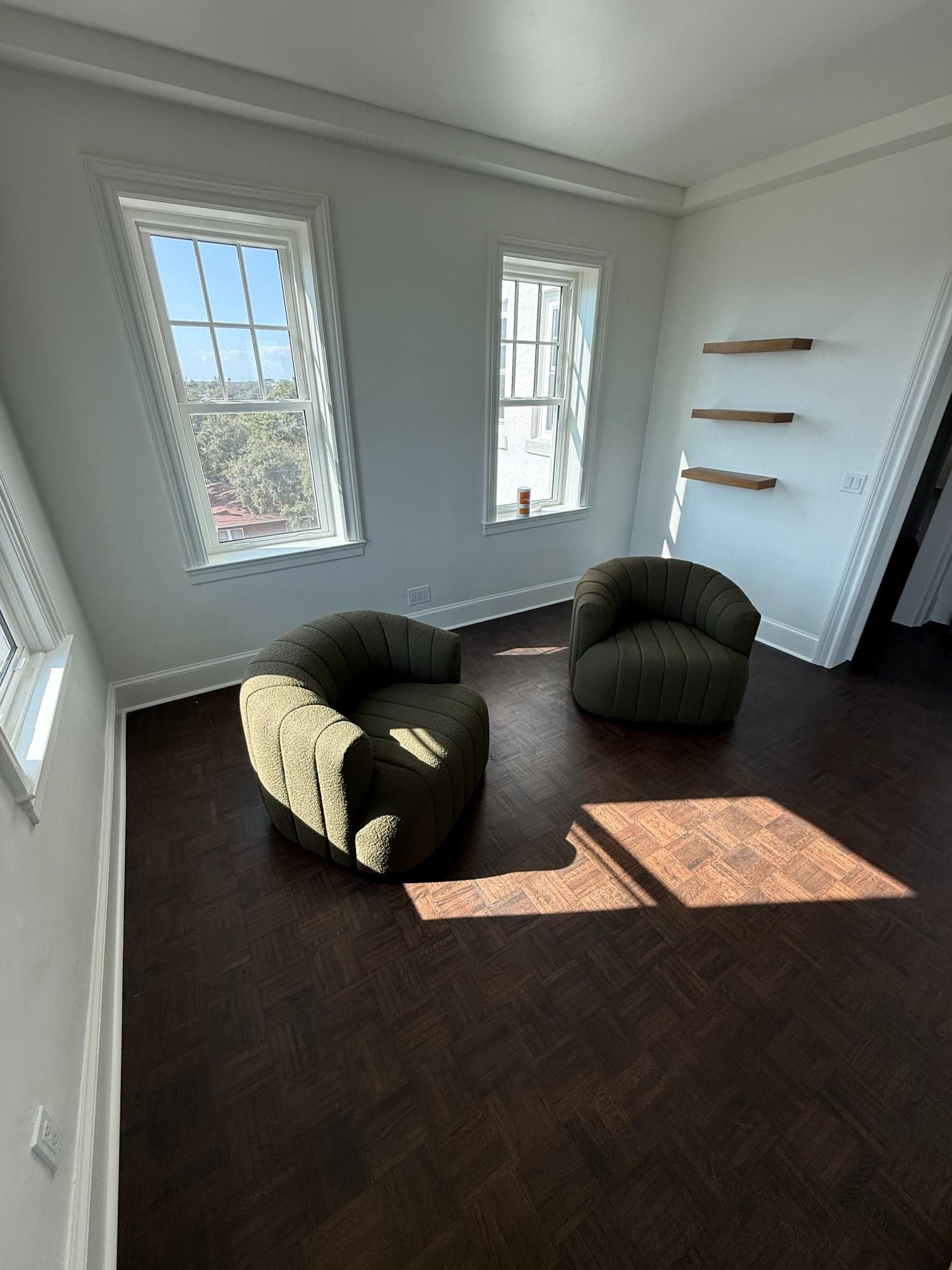 Two green armchairs in a room with hardwood floors, two windows, and floating shelves.