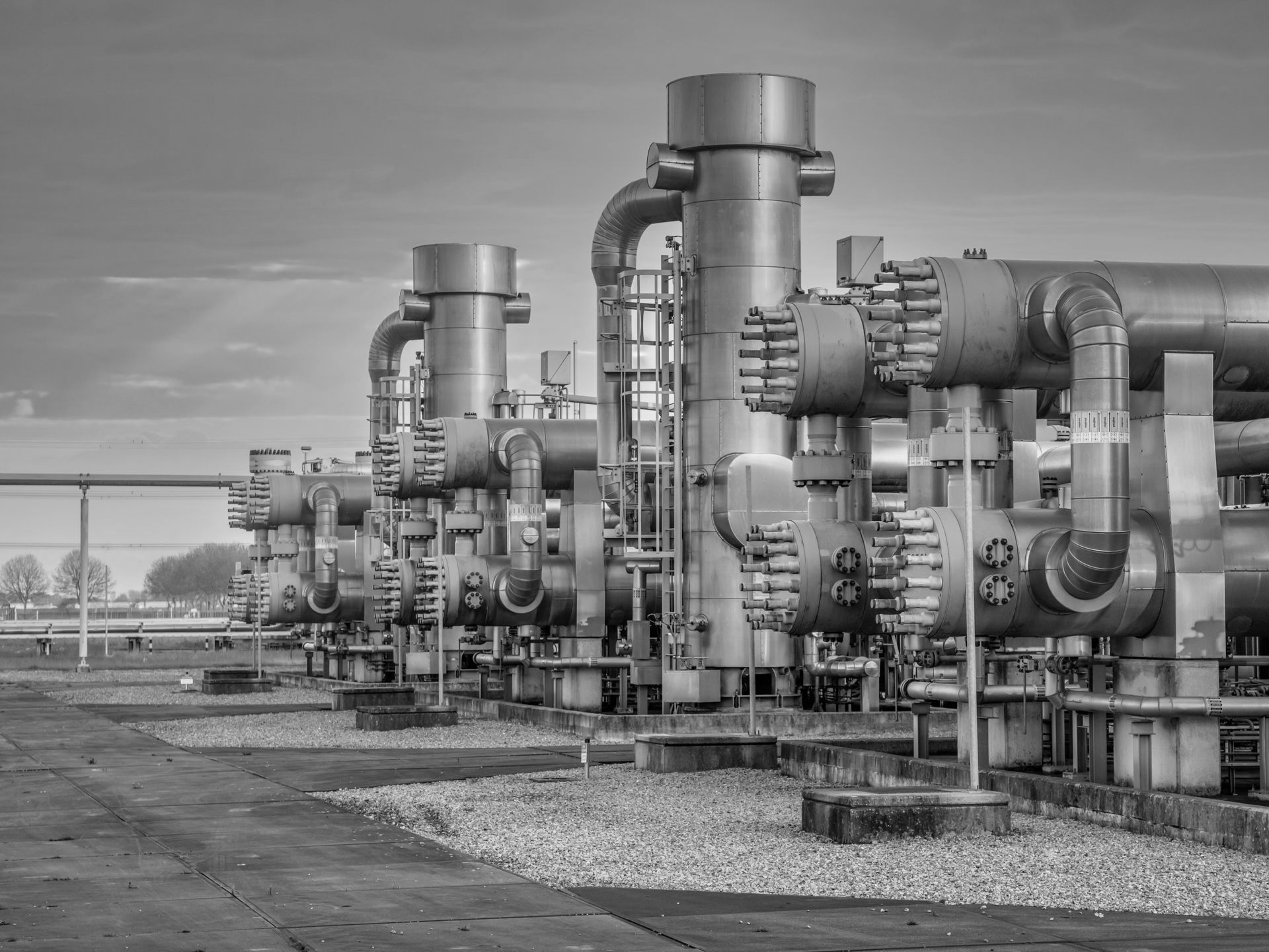 Industrial machinery with pipes and tanks, likely part of a gas or oil processing facility, outdoors on a gravel surface.