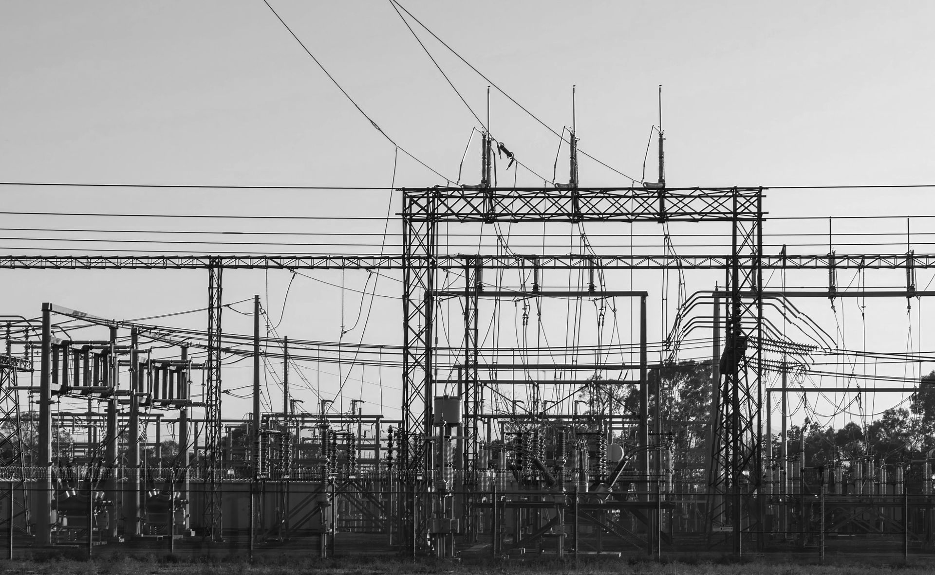 Black and white photo of a power substation with overhead wires, metal structures, and fencing against a clear sky.