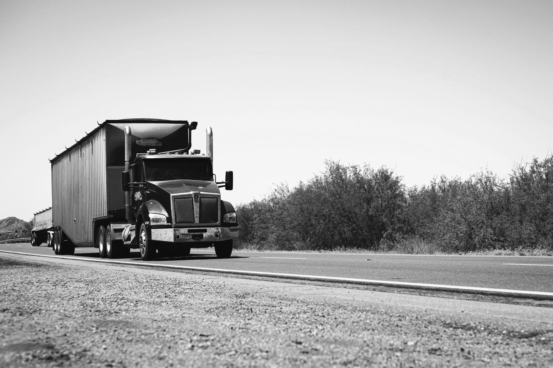 Semi-truck driving on a road, trees in the background, black and white.