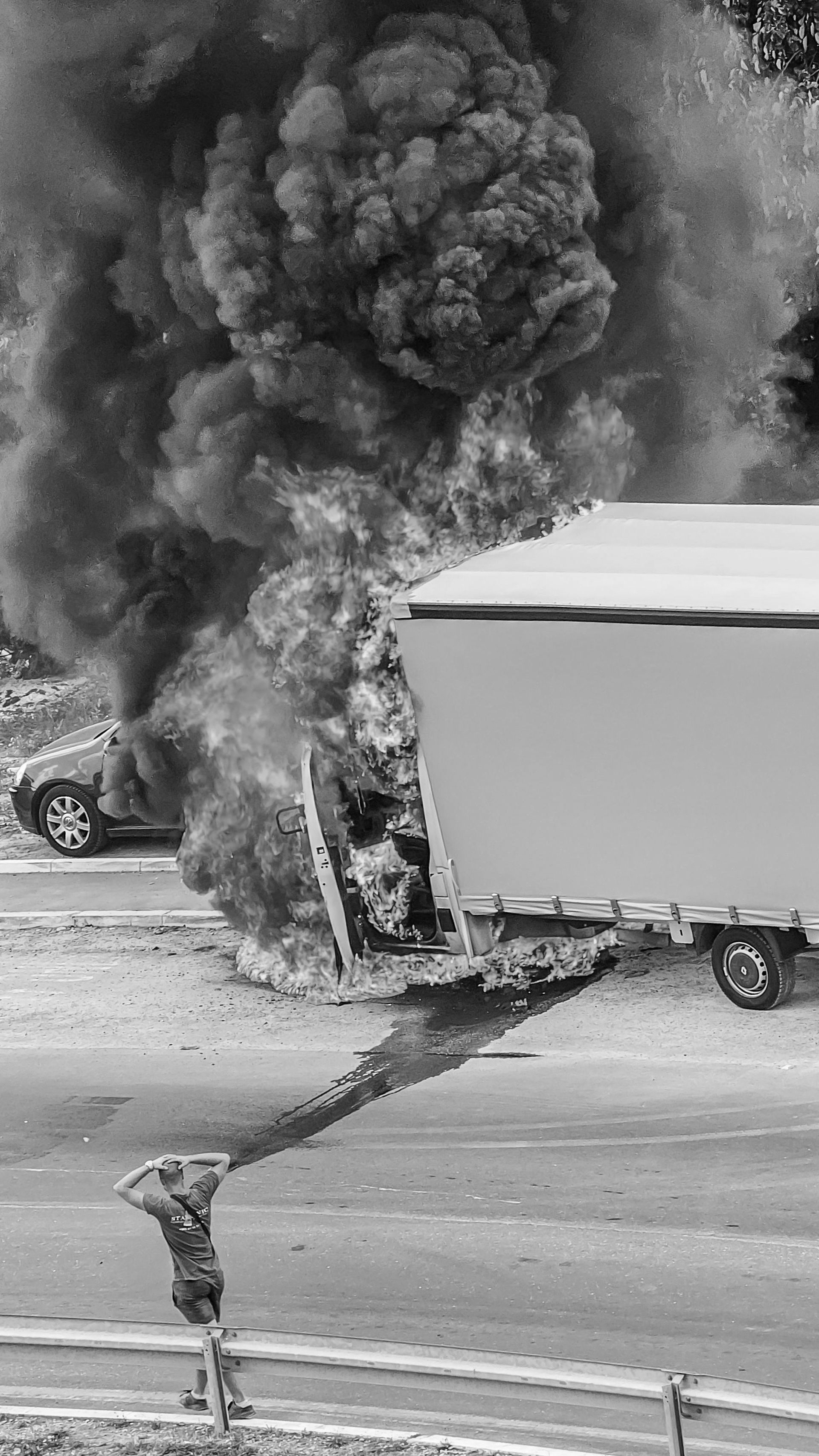 Truck engulfed in flames, thick black smoke billowing. Car also on fire. Fire is on a road.