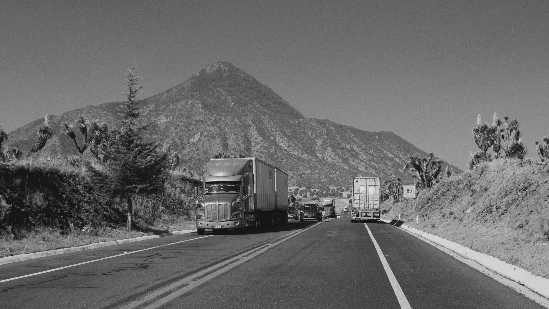 Black and white photo of a road with semi-trucks, leading to a mountain, set in a sunny landscape.