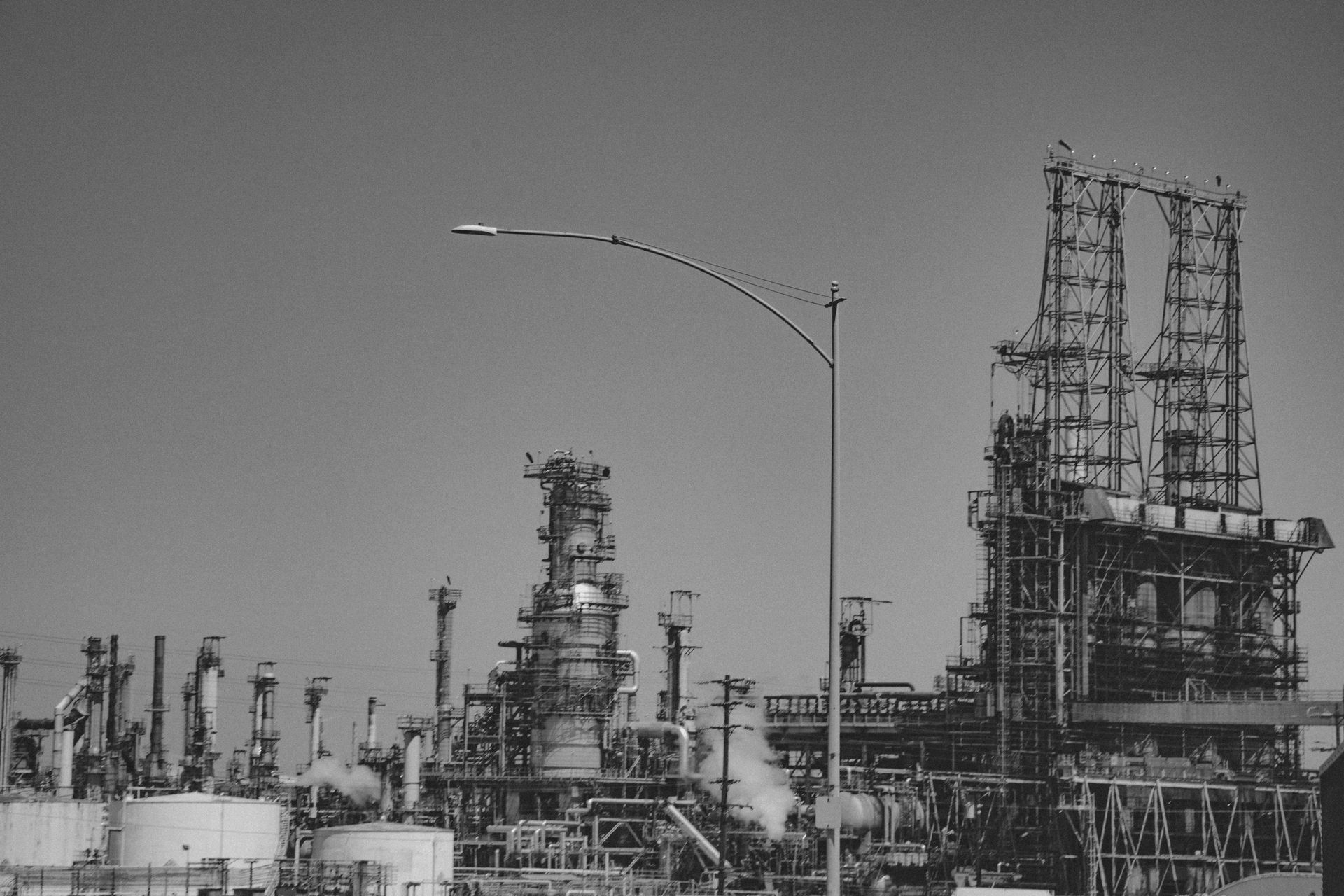Black and white photo of an oil refinery with tall structures, pipes, and tanks against a clear sky.