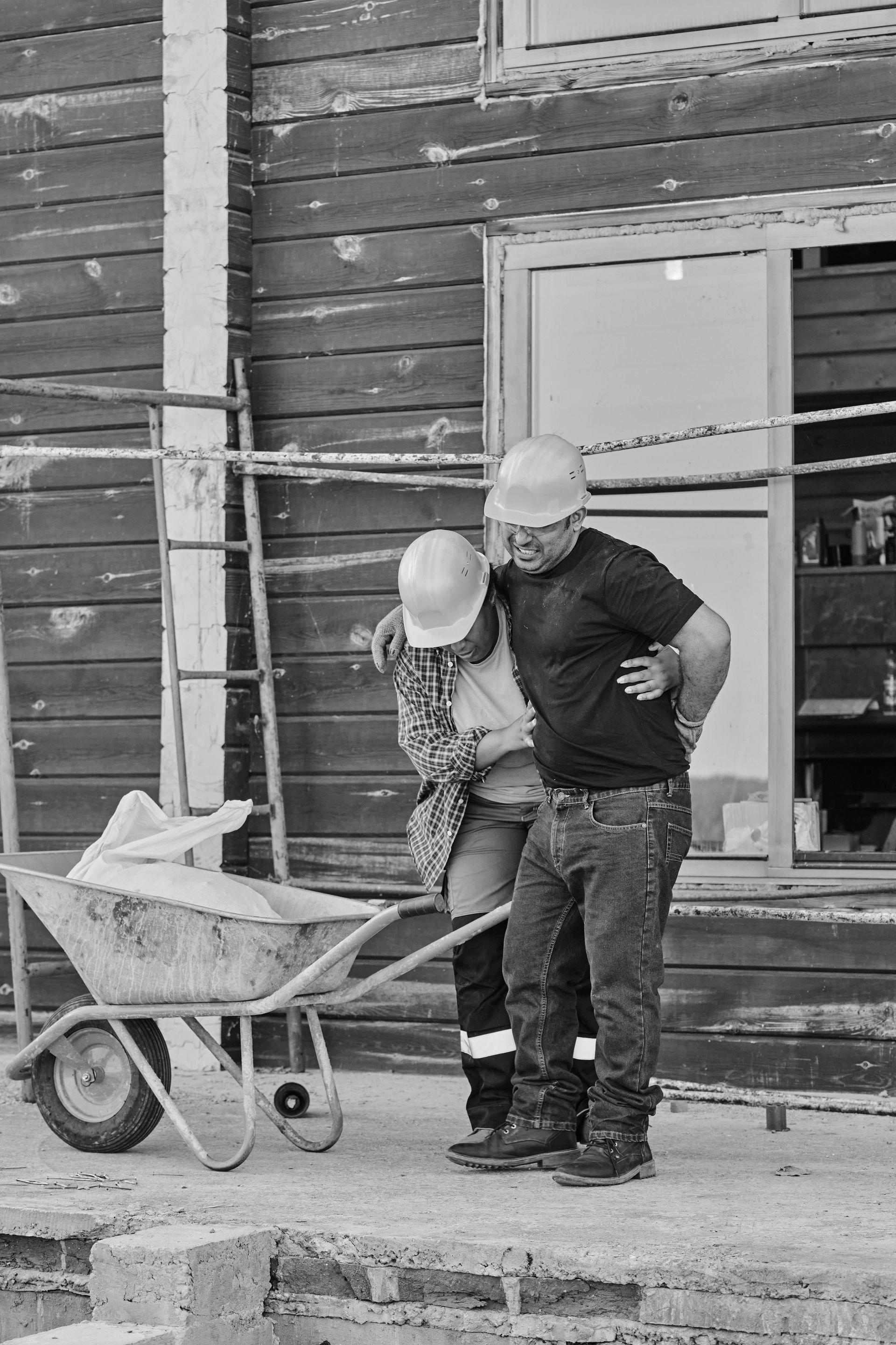 Two construction workers with hard hats near a wheelbarrow and wooden building. One worker appears to be guiding the other.