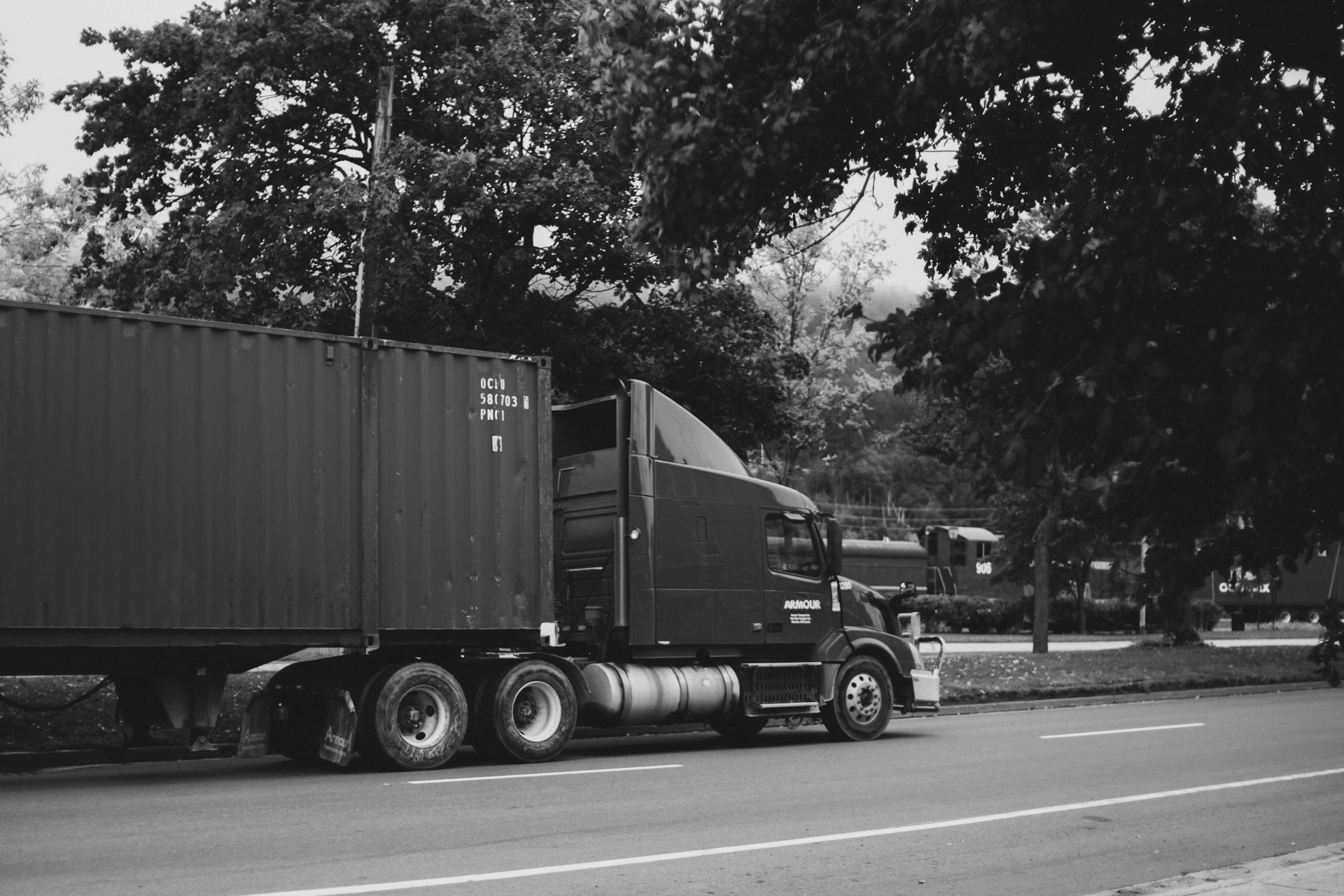 Black and white photo of a semi-truck parked on the side of a road, trees in the background.