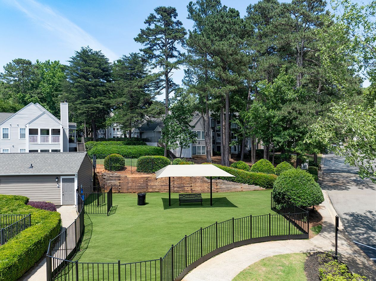 Dog park with green grass, black fence, and covered picnic area, set among trees and buildings at Station Heights in Alpharetta, GA.