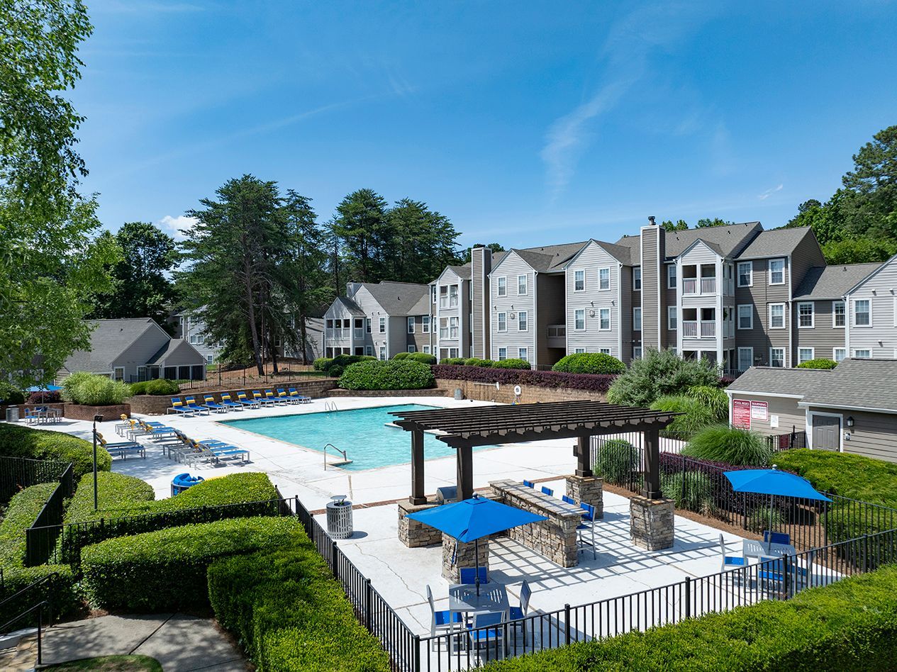 Apartment complex with pool. Buildings are gray with blue umbrellas, surrounded by greenery under a blue sky at Station Heights in Alpharetta, GA.