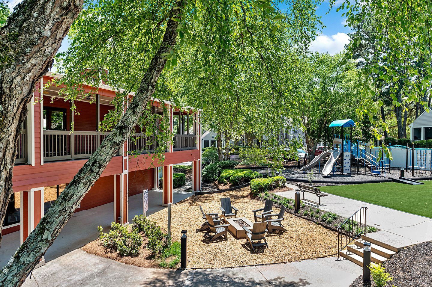 Red building with porch overlooking a courtyard with seating area, playground, and trees.