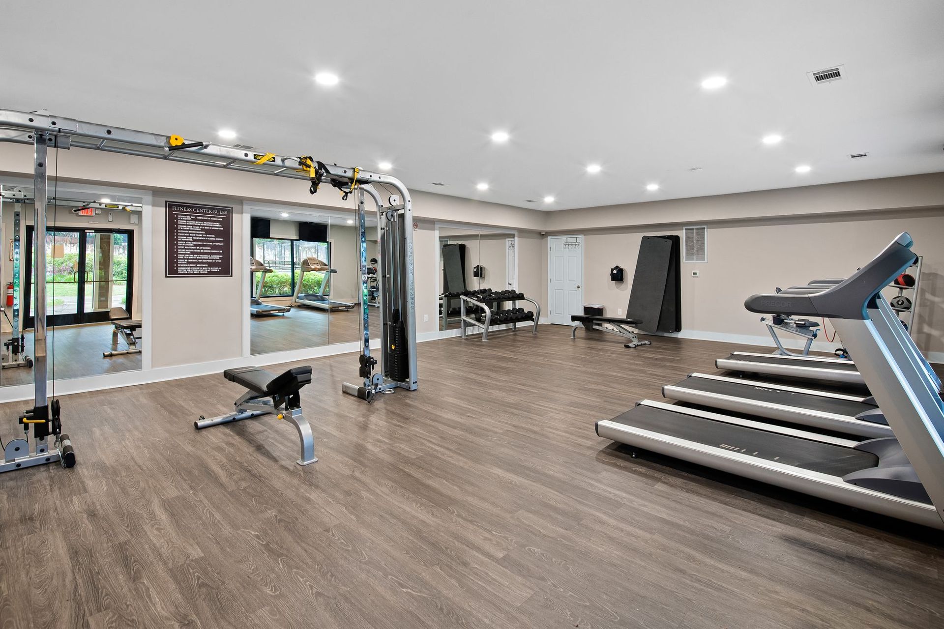 Gym with cardio machines, weight stations, and mirrors. Wood flooring, white walls, and bright lights.