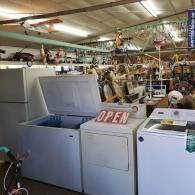 White kitchen appliances, including a freezer and two dryers, are displayed in a cluttered, warehouse-style retail store.
