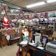 A thrift store interior with holiday tinsel draped over merchandise, including toys, a Santa figure, and office equipment.