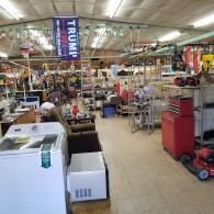 Interior of a cluttered shop with household appliances, tools, and a large Trump 2024 campaign flag hanging from the ceiling.