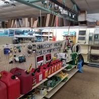 A retail hardware store aisle with red gas cans on a shelf, pegboard displays, and lumber stored on overhead racks.