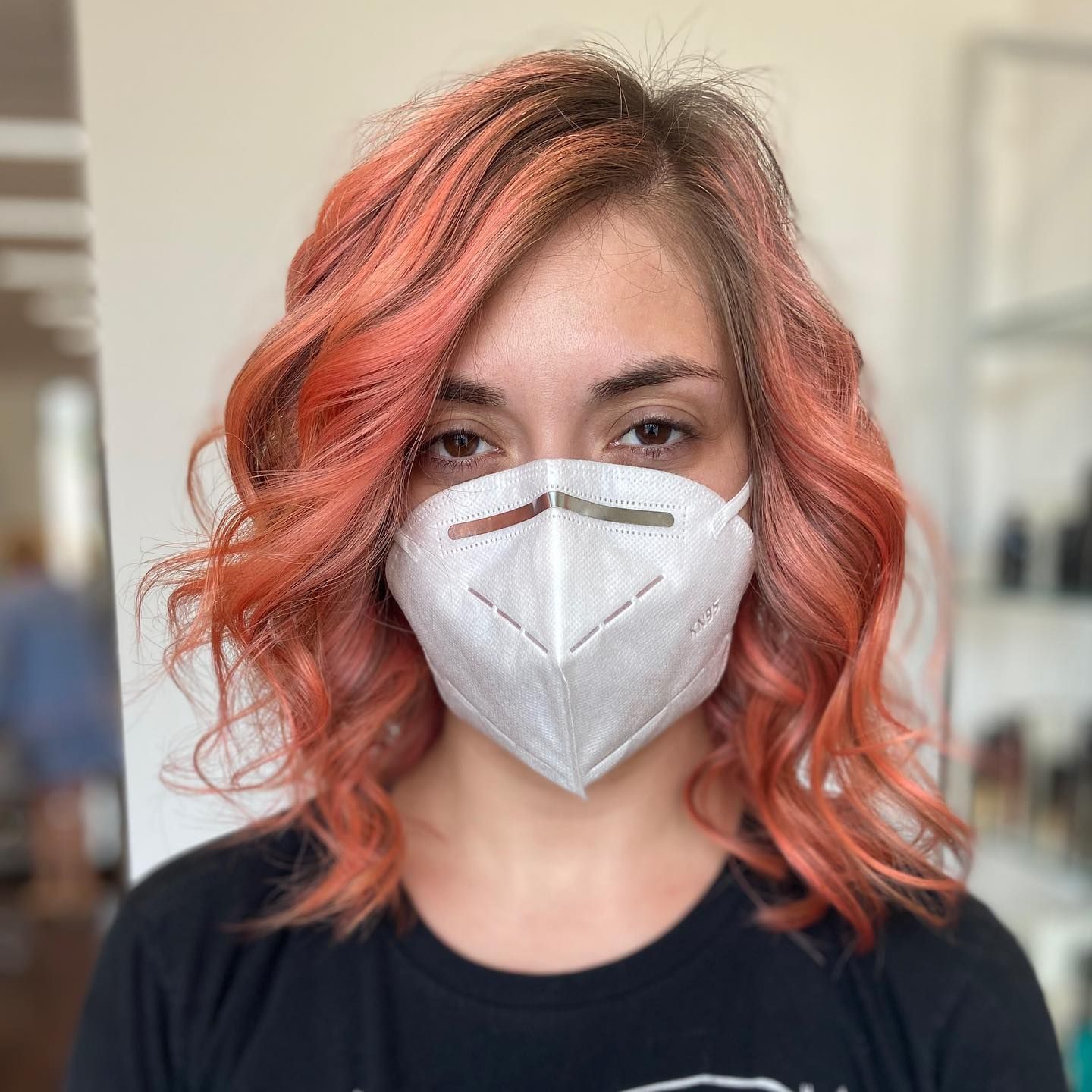 Woman with reddish-orange wavy hair wearing a white mask; indoors.