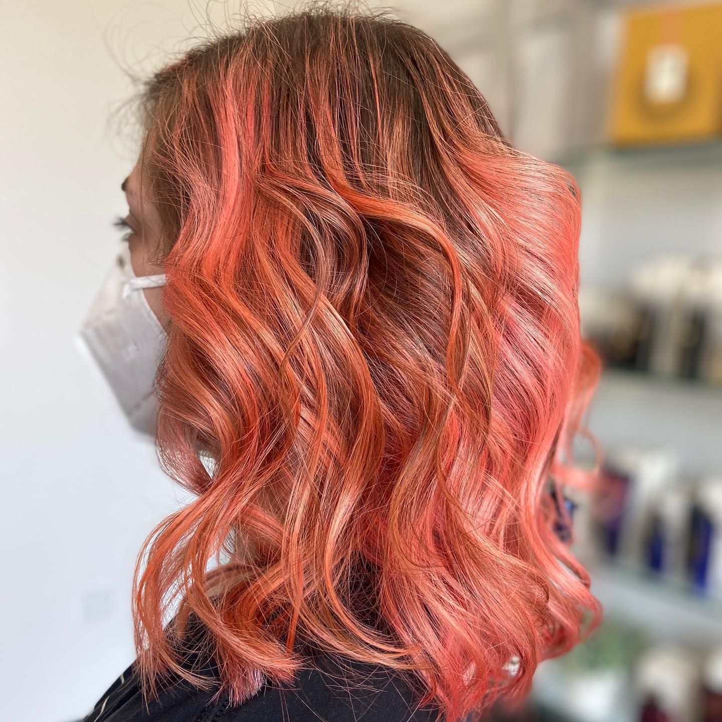 Woman with wavy, bright orange hair, wearing a white face mask in a salon.