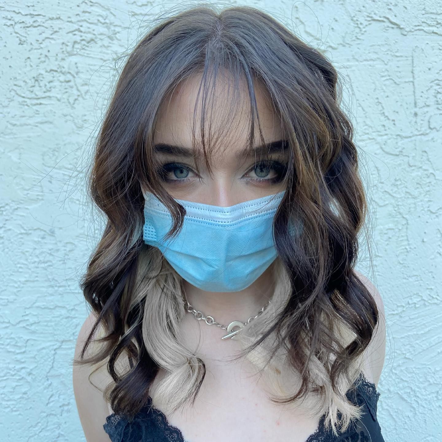 Woman wearing a blue face mask, wavy brown hair with blonde streaks, wearing a black top.