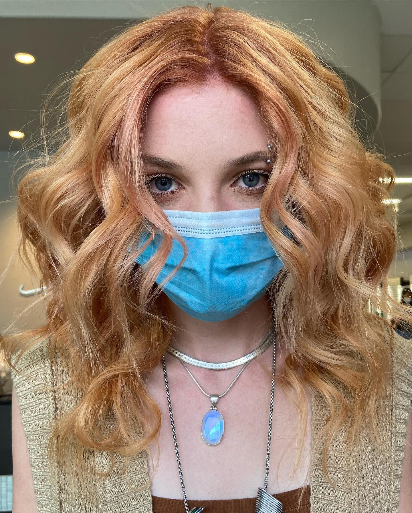 Woman with wavy red hair wearing a blue face mask and layered necklaces.
