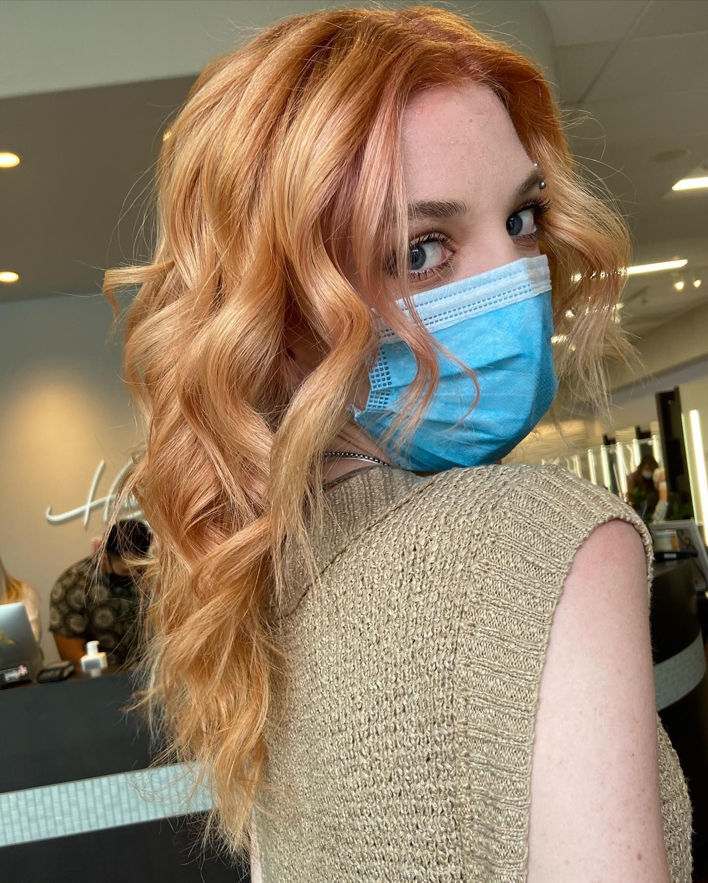 Woman with wavy, reddish-blonde hair wearing a blue face mask. She is in a salon, looking over her shoulder.