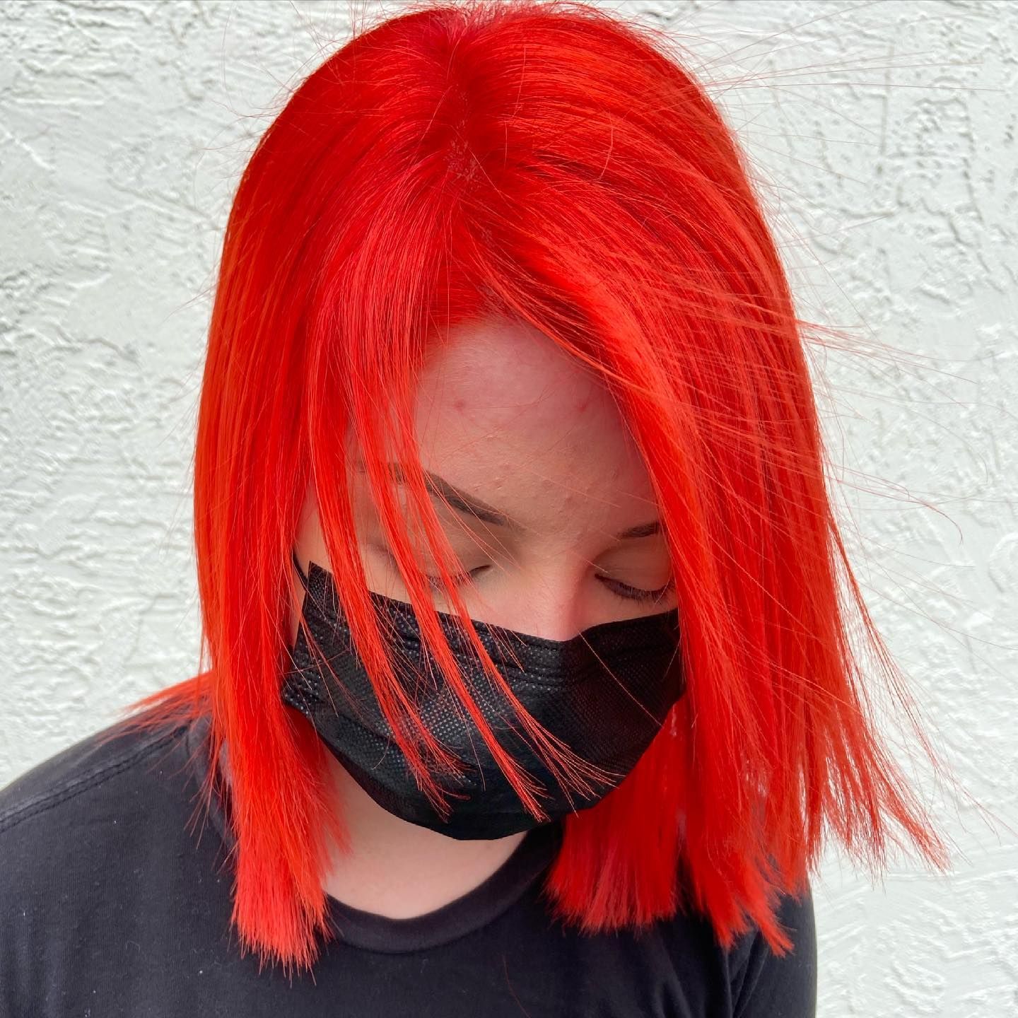 Person with bright red, chin-length hair, wearing a black face mask and a black shirt, eyes closed.