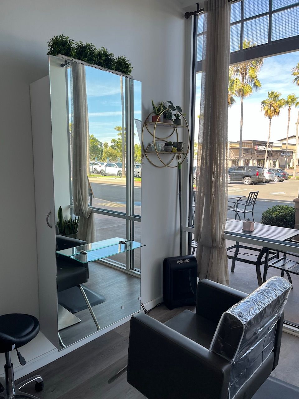 Hair salon interior with large mirror, black chair, and window view of palm trees and street.