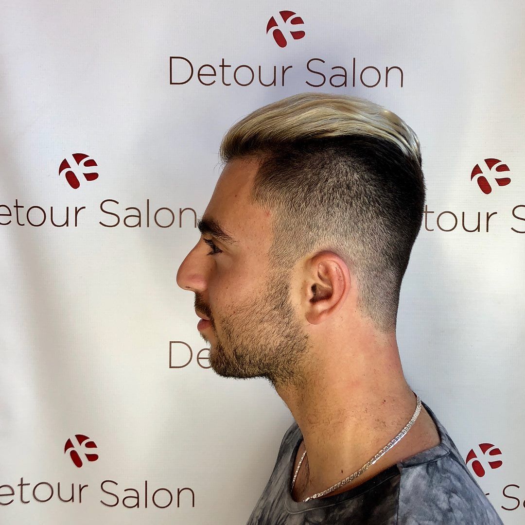 Man with a fade haircut, partially dyed blonde, in front of a salon backdrop.