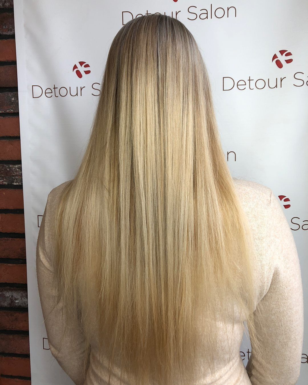 Woman with long, straight, blonde hair.  Wearing a light-colored top.  Standing in front of a salon backdrop.