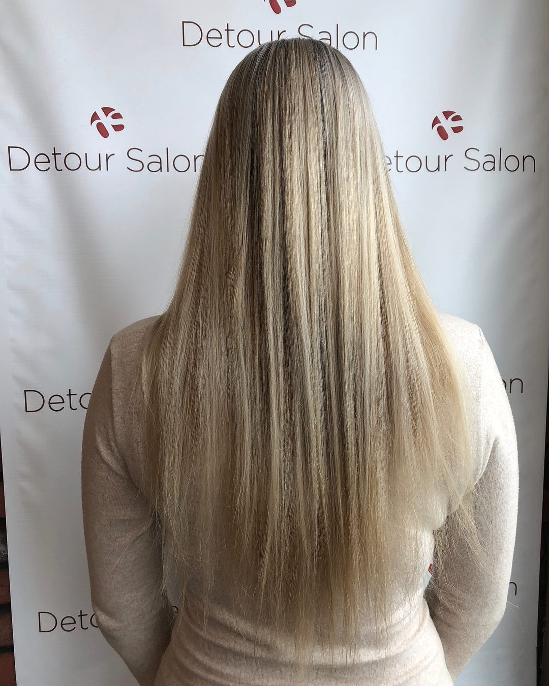 Blonde, straight hair, styled down the back, in front of a white wall with 