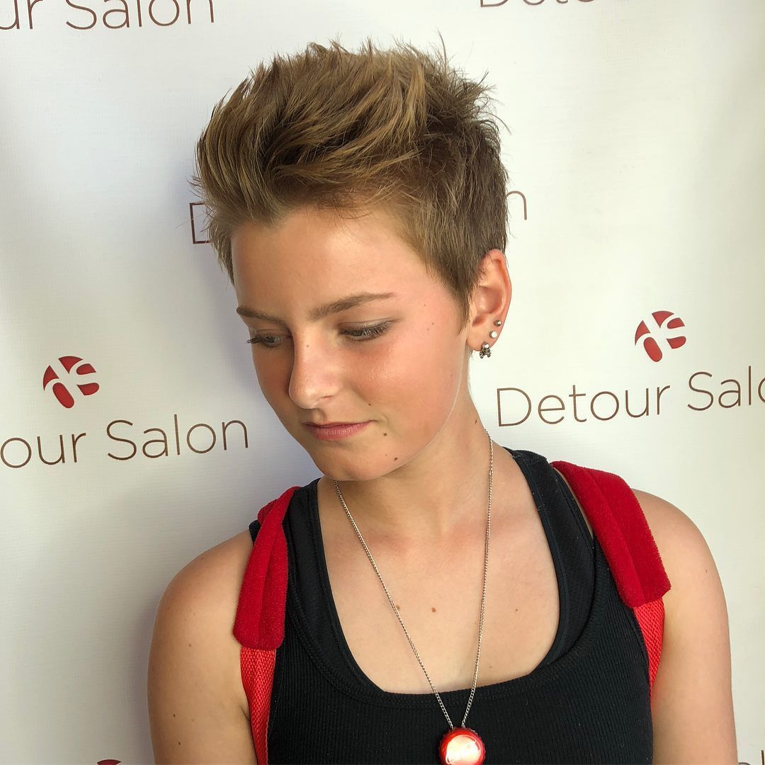 Person with short, spiky blonde hair, black tank top, red backpack straps. Leaning slightly, necklace.