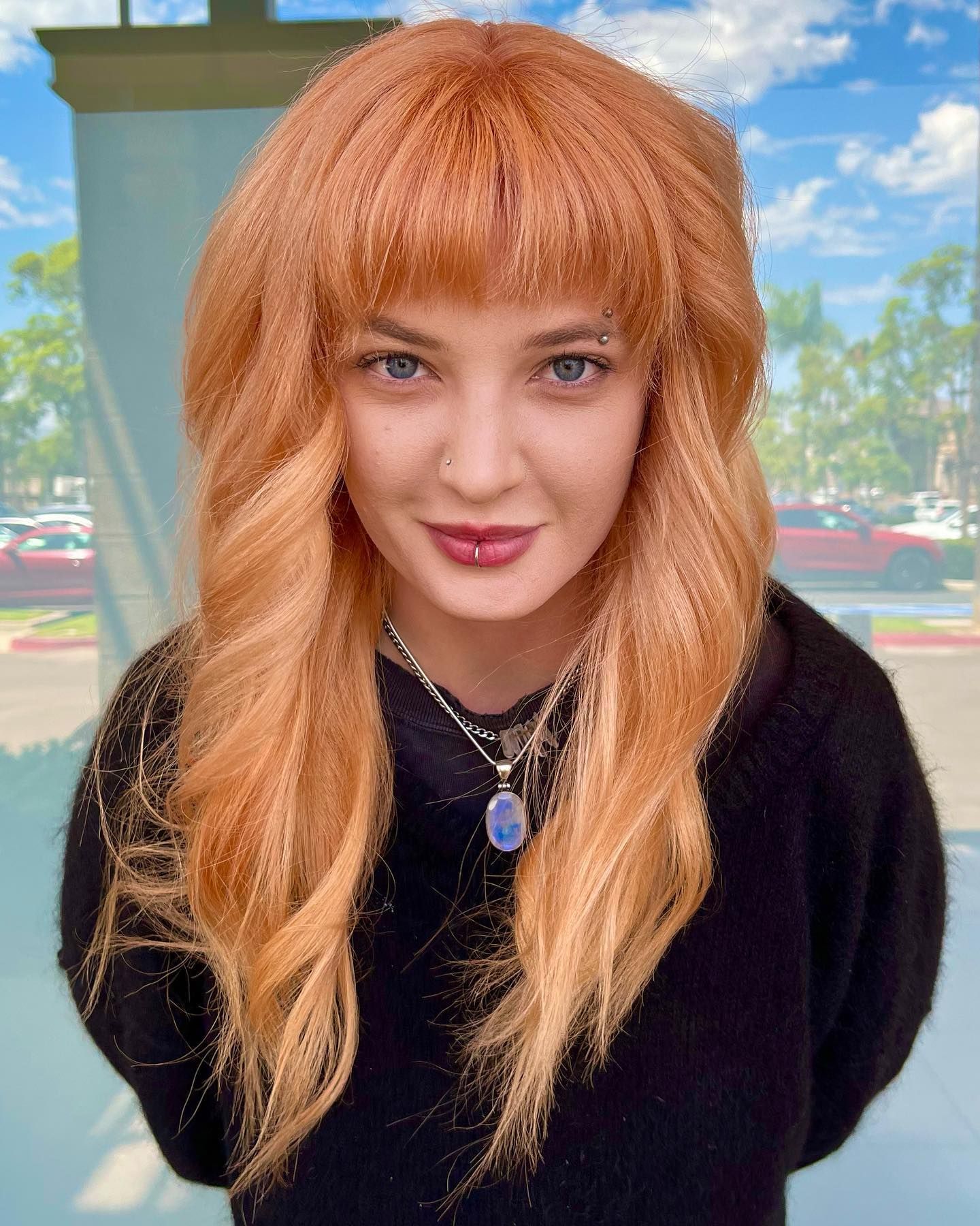 Woman with long, reddish-blonde wavy hair, bangs, wearing a black top and a pendant necklace, smiling slightly.