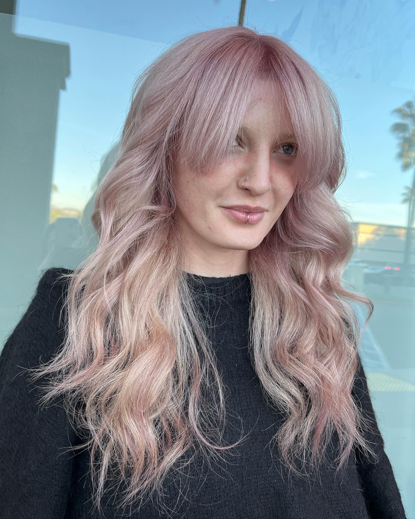 Woman with pink wavy hair, bangs, black sweater, smiling in front of a window.