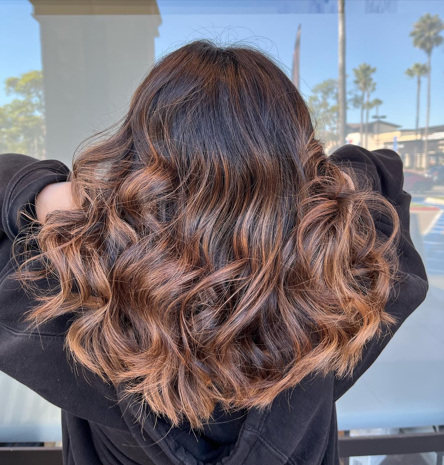 Woman with dark brown hair styled with brown highlights and loose curls, outside.