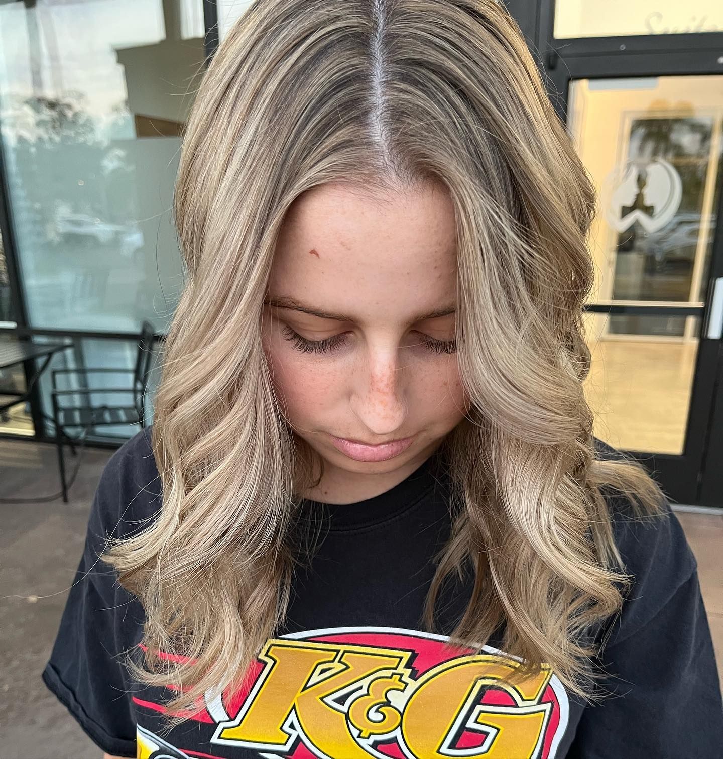 Woman with blonde, wavy hair and a black t-shirt, looking down. Outdoors, near a doorway.