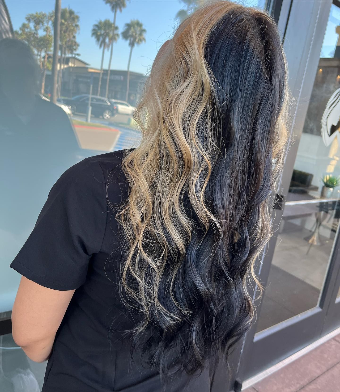 Person with wavy hair, split black and blonde. Leaning near a glass door in outdoor setting.