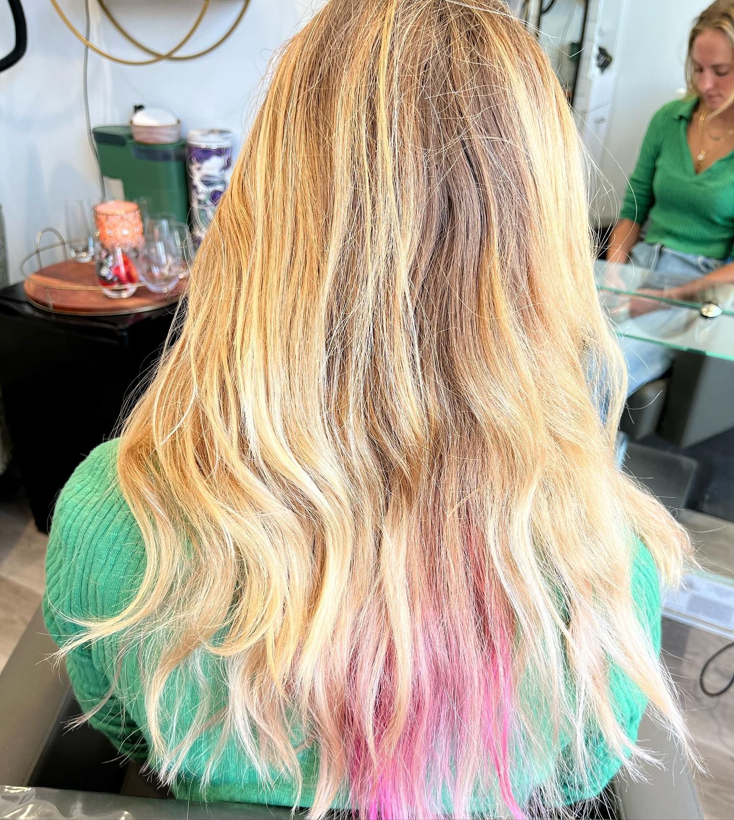 Woman with long blonde hair, pink tips, in a salon, wearing a green sweater.