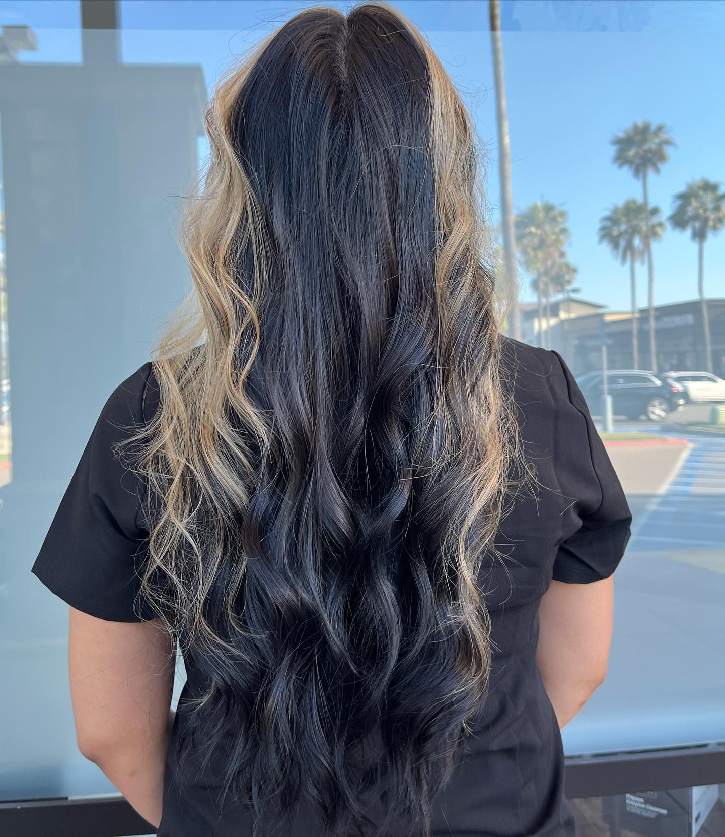 Woman with long, wavy black hair that transitions to blonde highlights. She wears a black shirt, outdoors with palm trees.