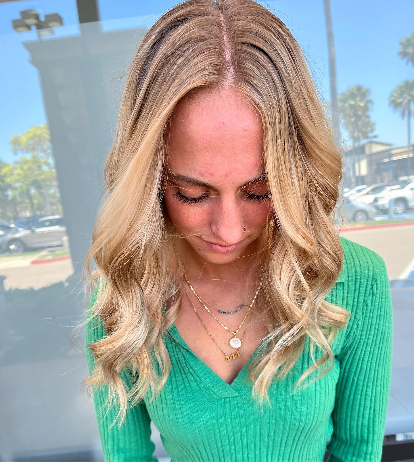 Woman with blonde wavy hair wearing a green top and necklaces, looking down, in front of a window.
